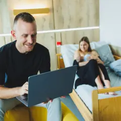 Mężczyzna z laptopem, korzystający z serwera VPN, siedzi obok łóżka, na którym odpoczywa kobieta z dzieckiem.