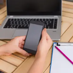 Ręce trzymają smartfon, obok leży laptop i notatnik z ołówkiem. Dowiedz się, jak rozpakować plik zip, by mieć dostęp do wszystkich danych.