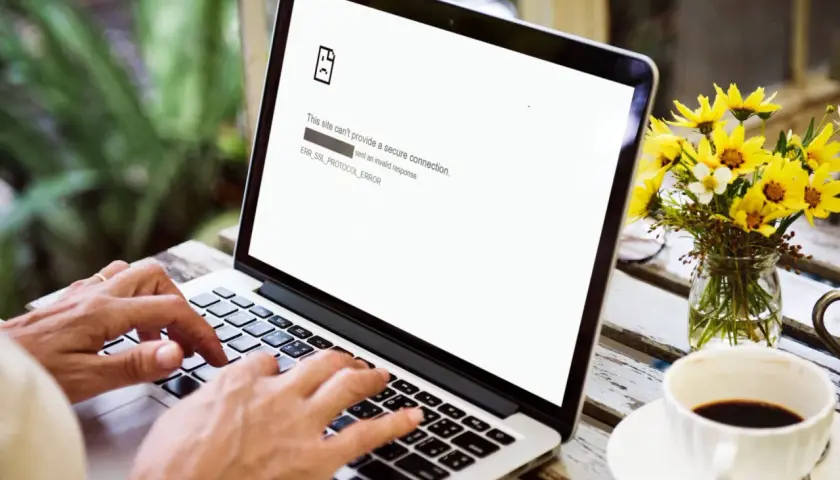 Osoba pisze na laptopie, na ekranie błąd ERR_SSL_PROTOCOL_ERROR. Obok kawa i kwiaty.