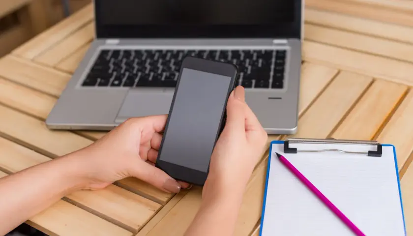 Ręce trzymają smartfon, obok leży laptop i notatnik z ołówkiem. Dowiedz się, jak rozpakować plik zip, by mieć dostęp do wszystkich danych.